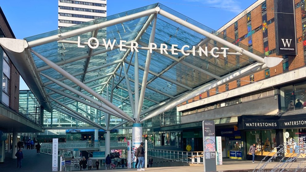 Entrance to Lower Precinct shopping area with glass canopy, people walking, and Waterstones bookstore visible on the right