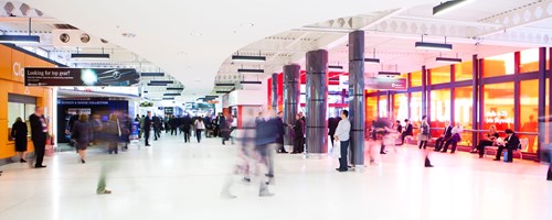 The image shows a corridor of the NEC in Birmingham. People in business attire are walking and sitting, in a brightly coloured environment.