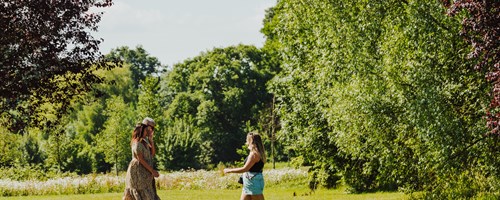 woman in shorts walking through park past another man and woman walking on the path in the park. sun is shining and it's very green.