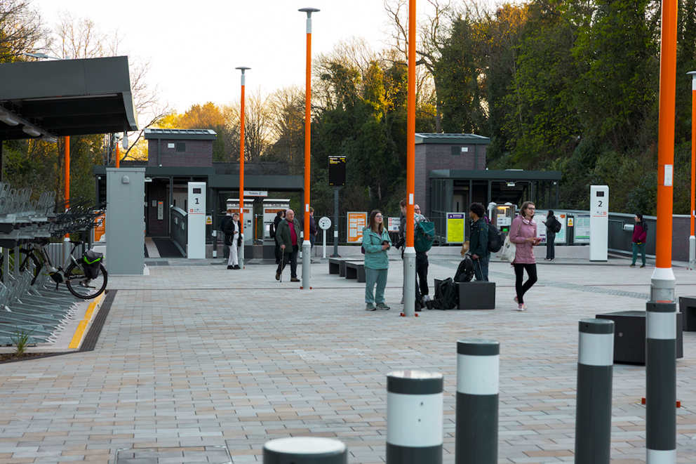 An image taken outside the front of Moseley Village railway station. There are people walking around the area, with the entrance to the station at the back and bike racks to the left.